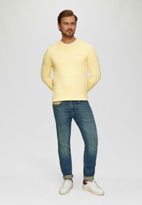 Gelber Pullover mit Rundhalsausschnitt, kombiniert mit blauen Jeans und weißen Sneakers. Der Pullover hat eine weiche Textur; die Jeans sind leicht zerstört.