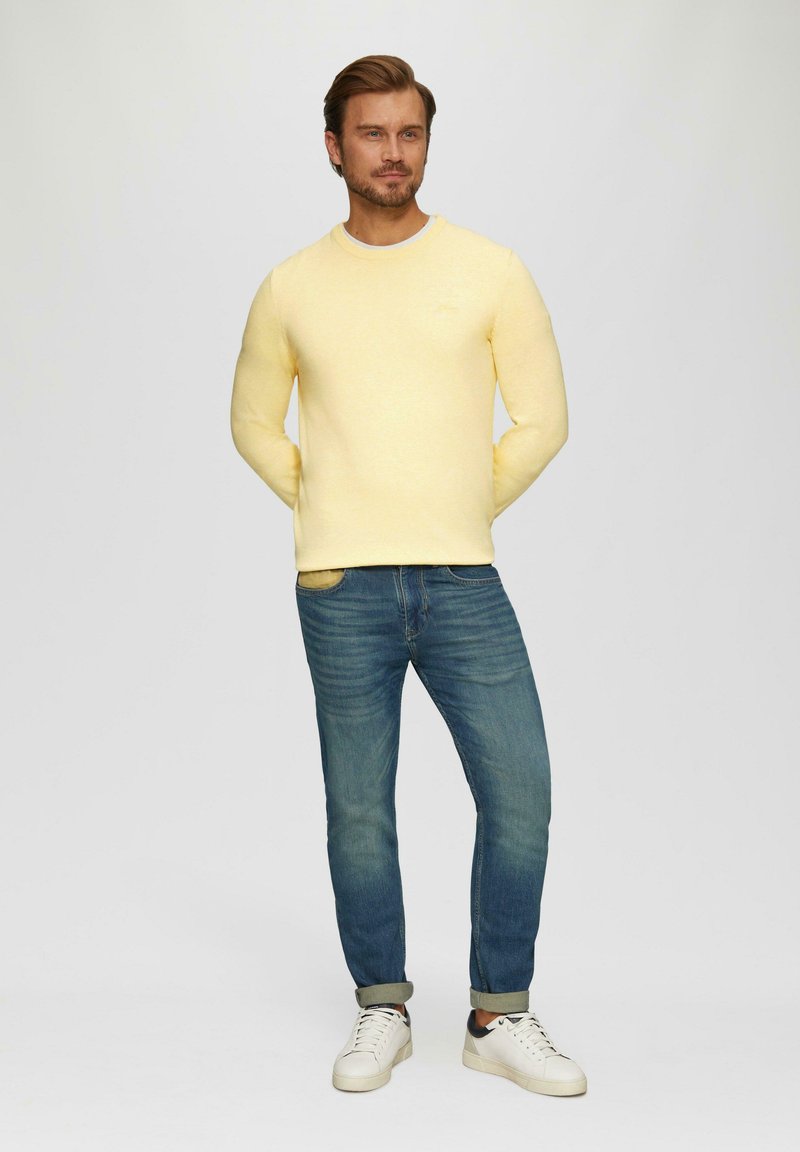 Gelber Pullover mit Rundhalsausschnitt, kombiniert mit blauen Jeans und weißen Sneakers. Der Pullover hat eine weiche Textur; die Jeans sind leicht zerstört.
