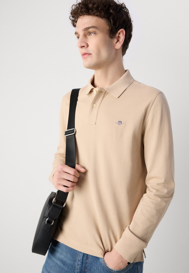 Beige Langarm-Polohemd mit Kragen, das ein kleines gesticktes Logo zeigt. Das Modell hält eine schwarze Umhängetasche mit einem Riemen.