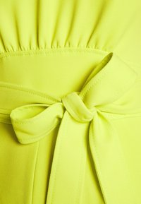 Tissu jaune brillant avec une large ceinture nouée en un joli nœud et des points froncés au-dessus de la taille.