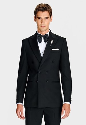 Czarna dwurzędowa marynarka od smokingu z satynowymi klapami, biała koszula formalna, czarna mucha, kieszeń na biodrze z białą poszetką oraz detal przypinki na klapie.