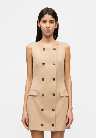 Robe beige sans manches avec un design croisé à six boutons marron, deux poches avant et une texture de tissu lisse.