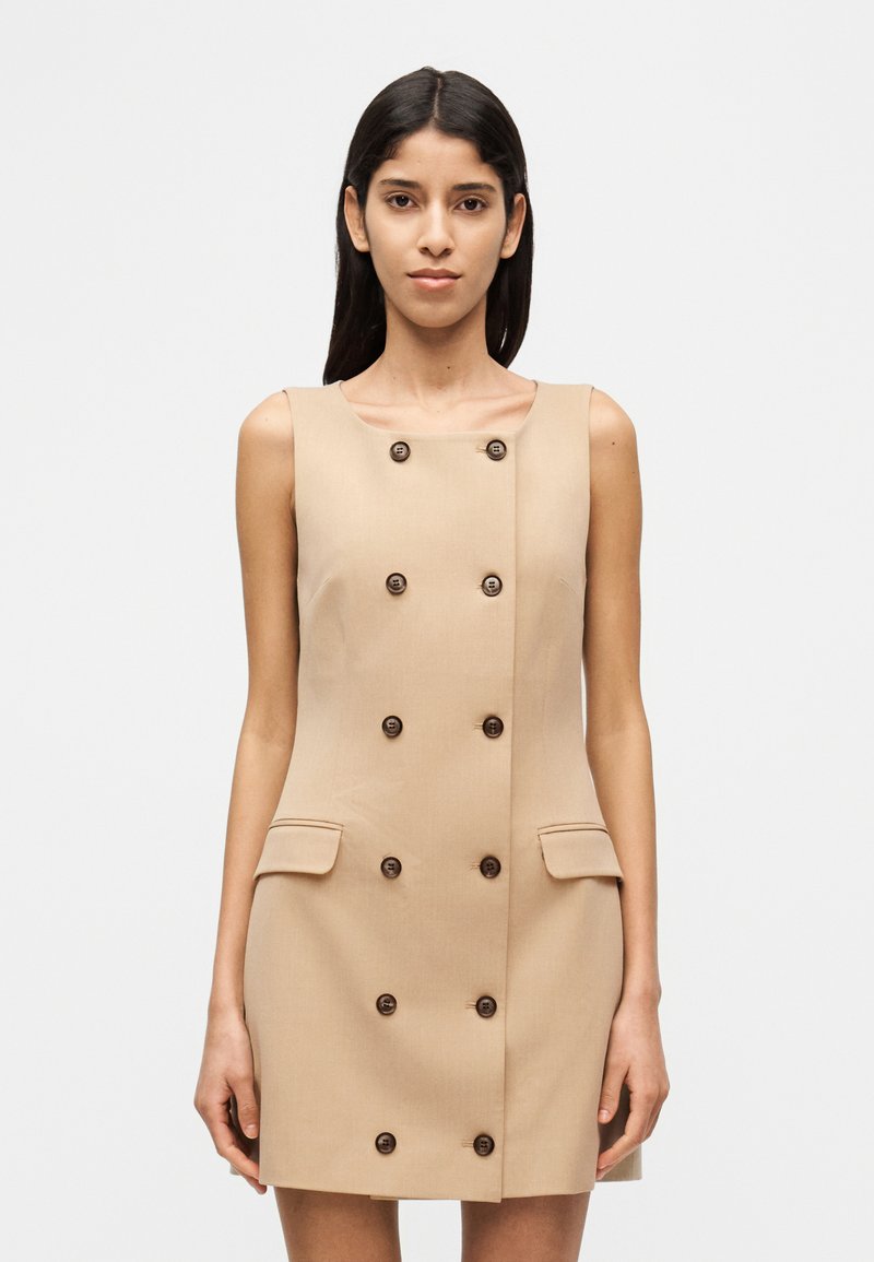 Beiges ärmelloses Kleid mit einem doppelt geknöpften Design, sechs braunen Knöpfen, zwei Taschen vorne und einer glatten Stoffstruktur.