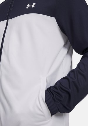 Wit- en marineblauwe jas met ritssluiting, hand in zijzak, met Under Armour-logo op de borst.