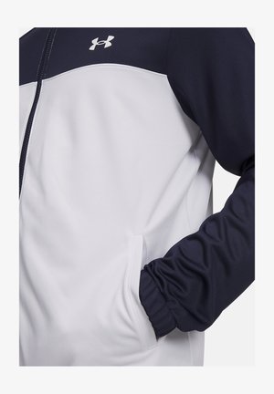 Wit- en marineblauwe jas met ritssluiting, hand in zijzak, met Under Armour-logo op de borst.