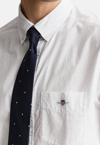 Homme portant une chemise blanche boutonnée avec un petit logo sur la poche et une cravate bleu marine à pois blancs.