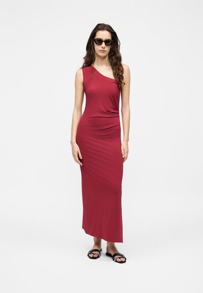 Γυναίκα με αμάνικο, κόκκινο μάξι φόρεμα με έναν ώμο, φοράει μαύρα σανδάλια και σκούρα γυαλιά ηλίου, στέκεται μπροστά σε λευκό φόντο.