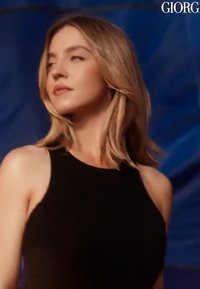 Femme blonde aux cheveux mi-longs portant un haut noir sans manches, regardant légèrement vers la gauche sur un fond bleu foncé.