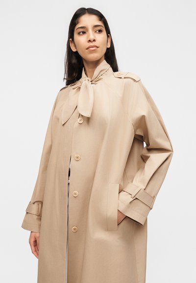 Γυναίκα που φοράει μπεζ παλτό τρεντσκότ με κλείσιμο με κουμπιά και δέσιμο στο λαιμό, ποζάρει με το ένα χέρι στην τσέπη μπροστά σε απλό φωτεινό φόντο.