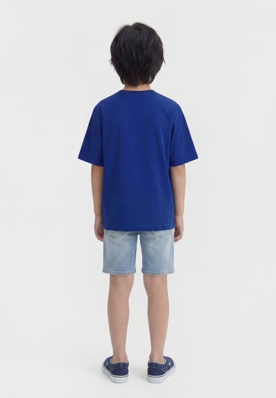 Παιδί που στέκεται με την πλάτη, φοράει απλό μπλε μπλουζάκι, ανοιχτόχρωμα τζιν σορτς και μπλε παπούτσια χωρίς κορδόνια πάνω σε λευκό φόντο.