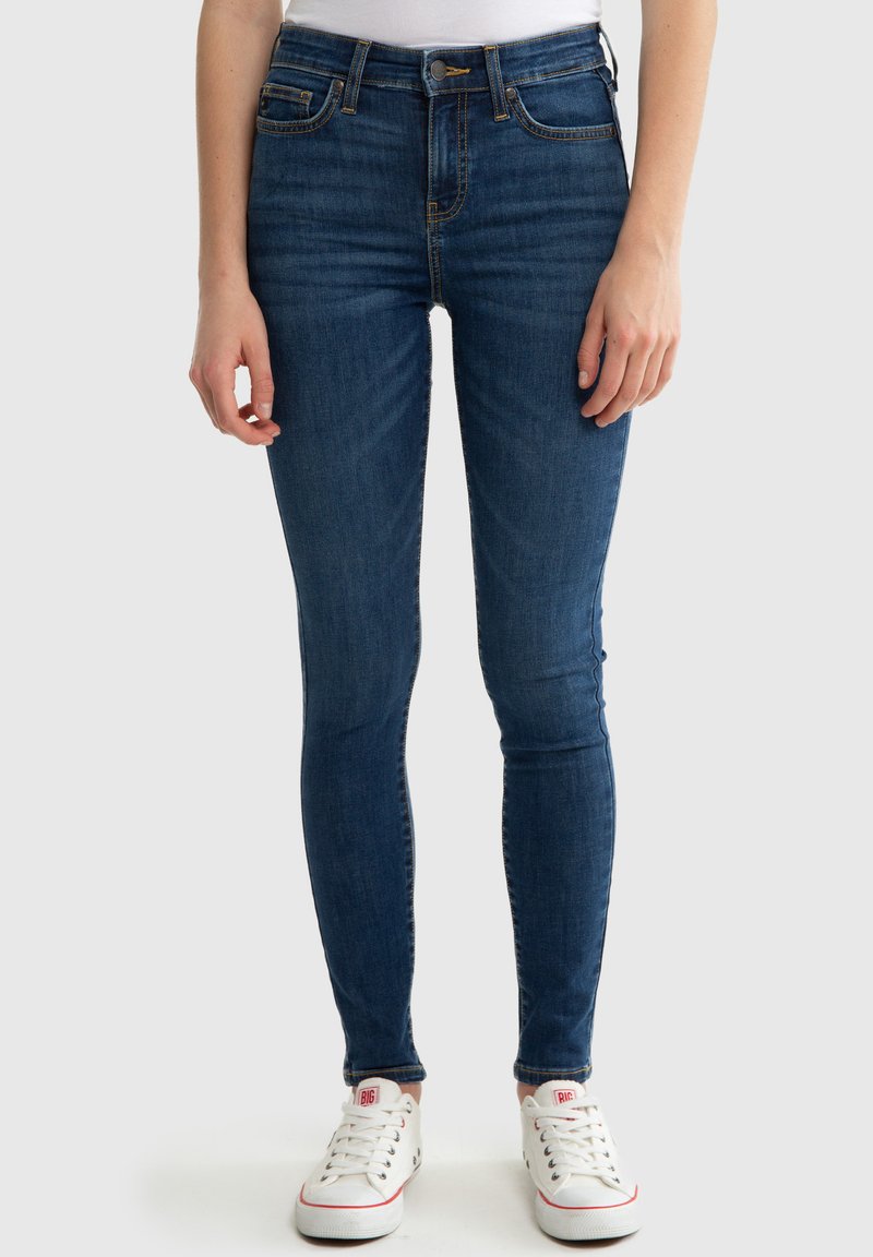 Jeans ajustados de mezclilla azul oscuro, con cintura alta, diseño de cinco bolsillos y costuras visibles. Combinados con zapatillas blancas.