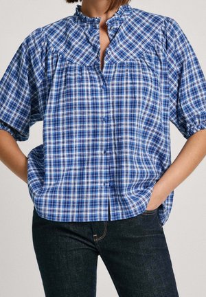 Vrouw draagt een blauw-wit geruit blouse met knopen en pofmouwen, en donkere denim jeans, handen in de zakken.