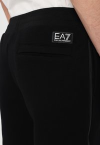 Čierne športové jogger nohavice s hladkým povrchom, s zadným vreckom a logom EA7 Emporio Armani. Detaily so zipsom na boku.