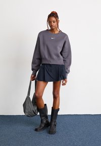 Joven con trenzas que lleva un sudadera gris de Nike, falda negra, botas negras y sostiene un bolso a cuadros, de pie sobre una alfombra azul.