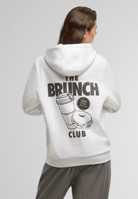 Biele mikina s veľkým čiernym grafickým motívom šálky kávy a pečiva s nápisom "KLUB BRUNCHU." Mäkko tkanina, voľný strih, kapucňa so sťahovacou šnúrou.