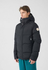 Zwarte pufferjack met een hoge kraag en capuchon. Voorzien van een zijzak met rits en een ronde logopatch op de bovenarm. Zachte textuur.