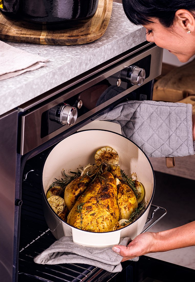 Biały ceramiczny garnek z pieczonym kurczakiem i ziołami, umieszczony w nowoczesnym piekarniku. Szary rękawiczkę kuchenną, stalowe pokrętła i blaty widoczne.