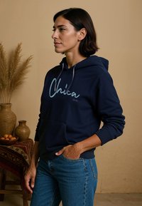 Marineblauwe hoodie met een voorzak, met "Chica" in lichtblauwe schreef. Geribde manchetten en onderrand, gecombineerd met blauwe jeans. Natuurlijke achtergrondelementen.