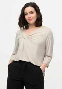 Kvinde med skulderlangt krøllet hår iført en beige bluse med tvistet knude og sorte bukser med hænderne i lommerne, stående mod en hvid baggrund.