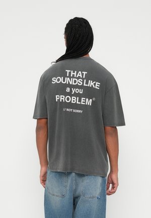 Muž s dlouhými zapletenými vlasy, který nosí tmavě šedé tričko s bílým textem "TO ZNÍ JAKO TVŮJ PROBLÉM // NENÍ MI TO LITOVAT" a světlé modré džíny.