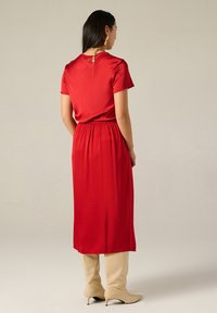 Abito rosso al ginocchio con maniche corte, vita elastica e dettagli in metallo dorato sul retro. Tessuto morbido, design semplice.