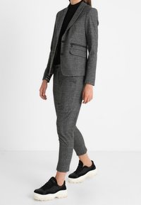 Szary teksturowany blezer z dopasowanymi, szytymi na miarę spodniami, obie części o wąskim kroju. Połączone z czarnymi trampkami z grubymi białymi podeszwami.