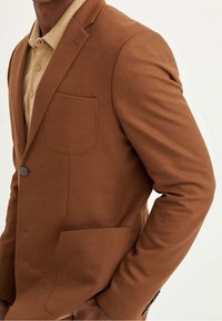 Bruine textuurblazer met een enkele borstzak, revers met inkeping en een sluiting met twee knopen aan de voorkant, gedragen over een beige overhemd met kraag.
