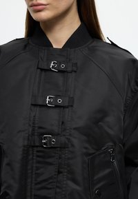 Μαύρο bomber μπουφάν με λεία υφή, ριμπ γιακά και πολλαπλές ασημένιες αγκράφες στο μπροστινό μέρος. Τσέπη με φερμουάρ στο πλάι.