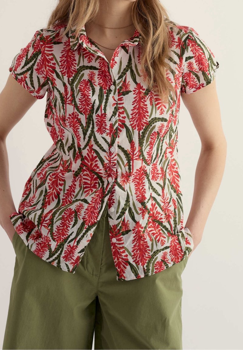 Femme portant une chemise à manches courtes avec un imprimé floral rouge et un pantalon vert olive, mains dans les poches.