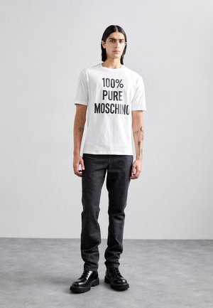T-shirt bianca in cotone con scritta nera in grassetto "100% PURE MOSCHINO", abbinata a jeans in denim scuri e stivali neri con lacci.