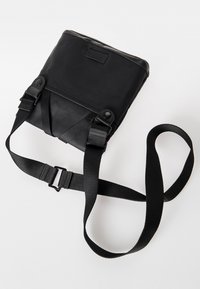 Black crossbody bag with adjustable nylon straps. Smooth texture, rectangular shape, and snap button details. Minimalist design.