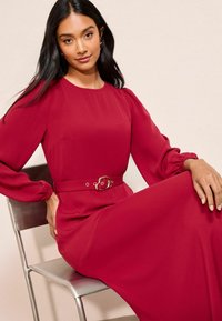 Rochie roșie cu mâneci lungi, având o croială lejeră, mâneci bufante și o curea cu accente aurii. Tesătură netedă cu o siluetă fluidă.