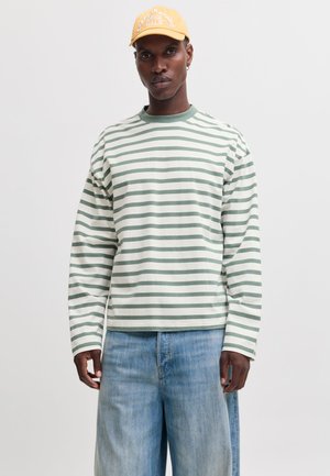 Homme portant une chemise à manches longues rayée verte et blanche, un jean bleu clair et une casquette jaune avec un texte blanc, debout devant un fond uni.