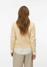 Žena s dlouhými hnědými vlasy v béžovém svetru, bílé košili a světle šedých kalhotách, stojící čelem k jednobarevnému bílému pozadí.