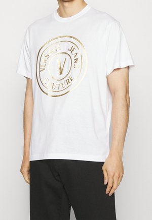 Mężczyzna w białej koszulce z krótkim rękawem z złotym logo Versace Jeans Couture i czarnych spodniach.