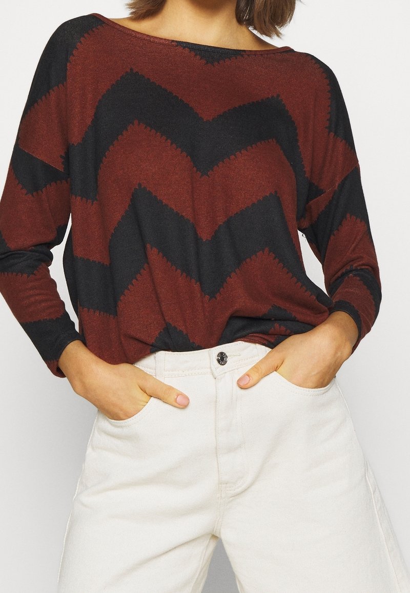 Długi rękaw z wzorem w zygzaki w rdzawoczerwonym i czarnym kolorze, wykonany z miękkiego materiału. Połączony z białymi spodniami o wysokim stanie.