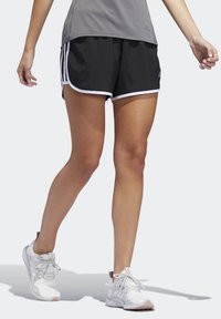 Pantalones cortos deportivos negros con ribete blanco, con un ajuste holgado y cortes laterales, combinados con zapatillas deportivas blancas y una camiseta gris.
