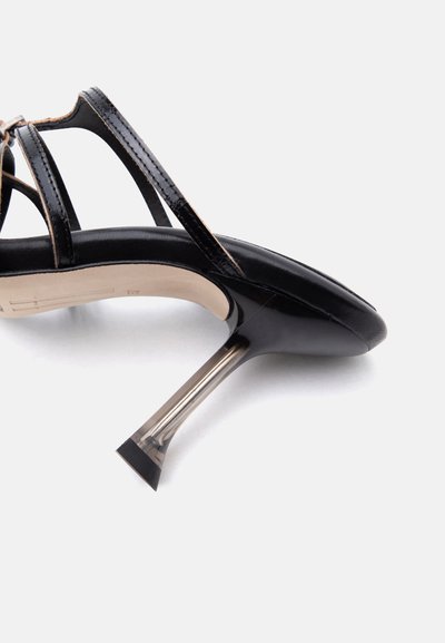 Μαύρο παπούτσι με τακούνι από δέρμα πατέντας, με λουράκια με κοψίματα και διαφανές φαρδύ στιλέτο τακούνι σε λευκό φόντο.