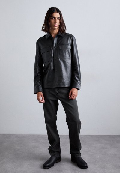 Young man with long hair wearing a black leather jacket, dark jeans, and black boots standing against a plain gray wall.