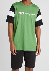 Camiseta verde de manga corta con mangas negras y detalles en blanco. Presenta el logo "Champion" en blanco en el pecho. Tejido suave de algodón.