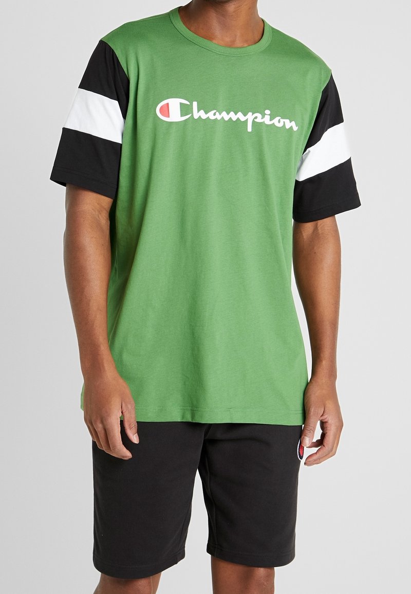 Camiseta verde de manga corta con mangas negras y detalles en blanco. Presenta el logo "Champion" en blanco en el pecho. Tejido suave de algodón.