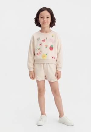 Παιδί που φοράει μπεζ φούτερ με πολύχρωμες εκτυπώσεις φρούτων και λουλουδιών, ταιριαστά σορτς με βολάν και λευκά αθλητικά παπούτσια, στέκεται μπροστά σε λευκό φόντο.