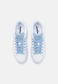 Zapatillas blancas con cordones azul claros y acentos de felpa azul en la lengüeta. Área del dedo del pie perforada, suela de goma y logotipo en la plantilla.