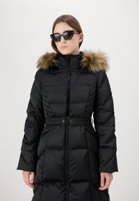 Juodas pūkinis paltas su dirbtinio kailio kapisonu, siuvinėtas dizainas, užtrauktuko uždarymas ir dirželis liemens srityje, su apvaliu sagties akcentu.