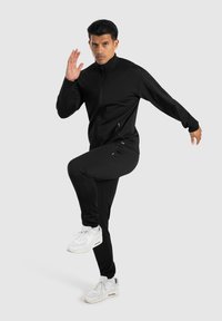 Zwarte sportieve trainingspak met een ritsjack en slim-fit broek, gecombineerd met witte sneakers; gladde stof en praktisch ontwerp.