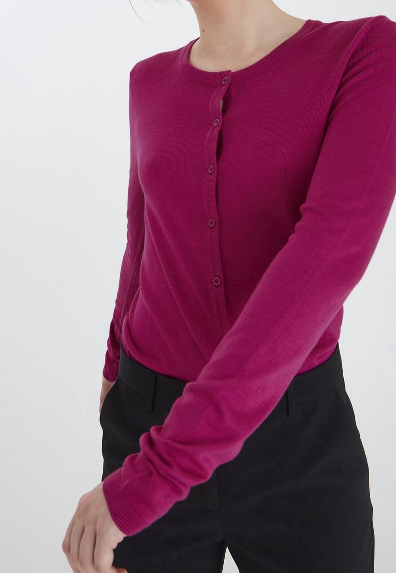 Przylegający magenta kardigan z długimi rękawami, okrągłym dekoltem i siedmioma guzikami z przodu; miękka tekstura; w zestawieniu z czarnymi spodniami.