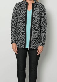 Donna che indossa una giacca zip-up leopardata nera e grigia sopra una maglietta azzurra a V e pantaloni neri, in piedi contro uno sfondo neutro.