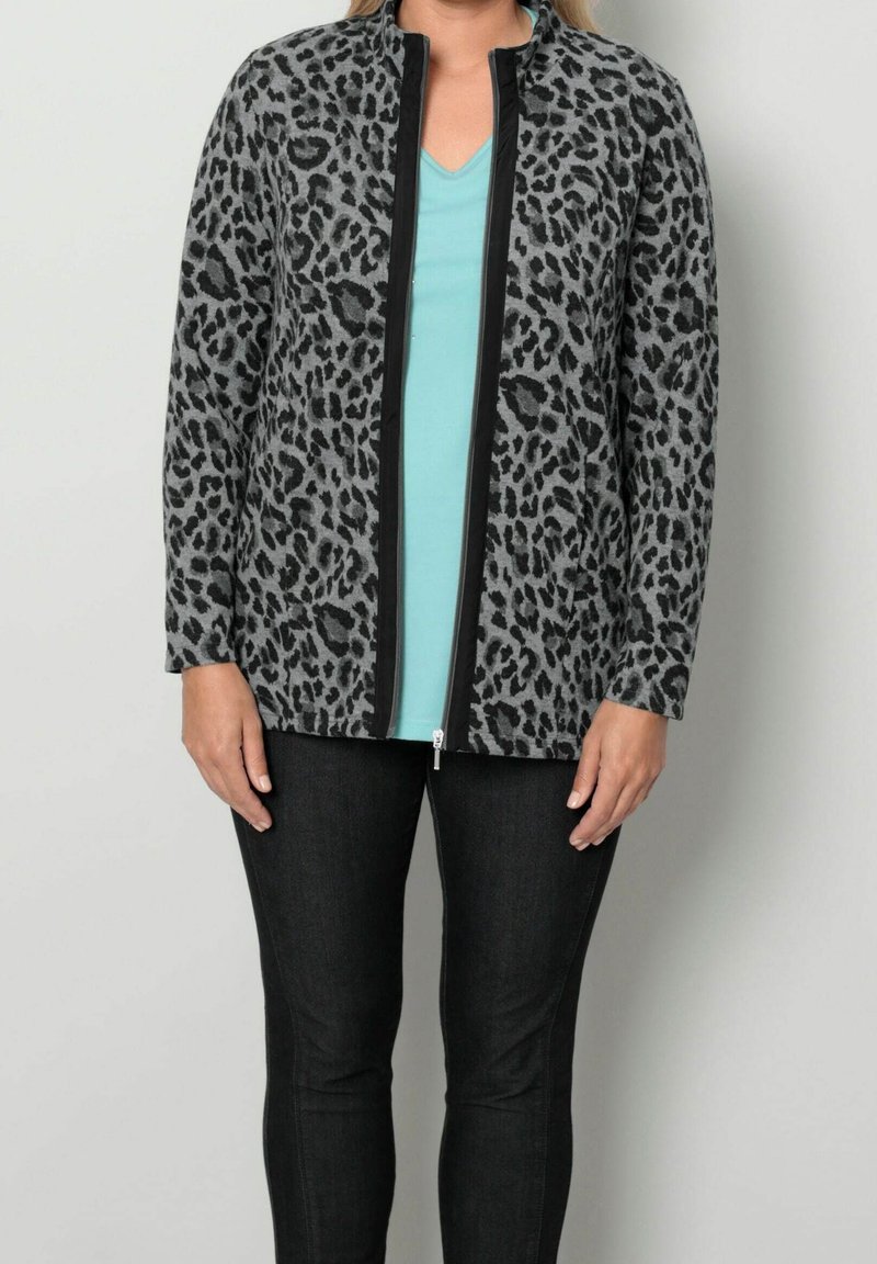 Donna che indossa una giacca zip-up leopardata nera e grigia sopra una maglietta azzurra a V e pantaloni neri, in piedi contro uno sfondo neutro.