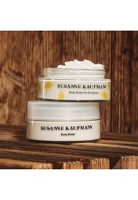 Des pots de beurre corporel empilés, présentant un design en verre dépoli. Le pot supérieur affiche un motif d'oiseau jaune. Étiquettes en texte noir sur les deux pots.