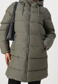 Abrigo acolchado verde oliva con capucha, que cuenta con una cremallera frontal, bolsillos laterales y un diseño acolchado con paneles cosidos.